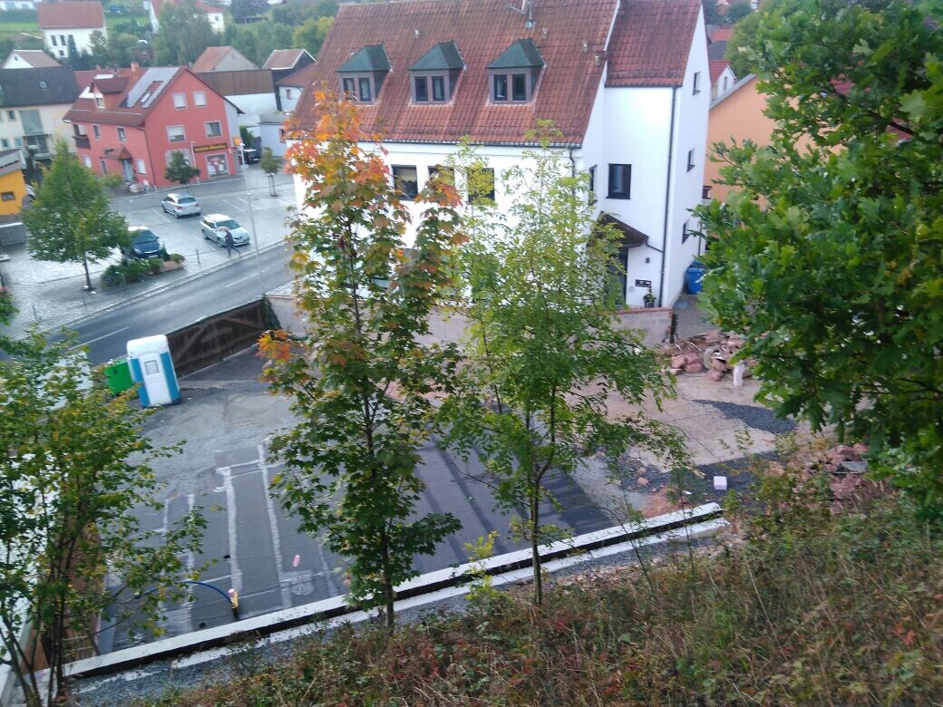 Blick von oben auf eine städtische Baustelle mit Porta-Potty, roten Dachhäusern und Bäumen.
