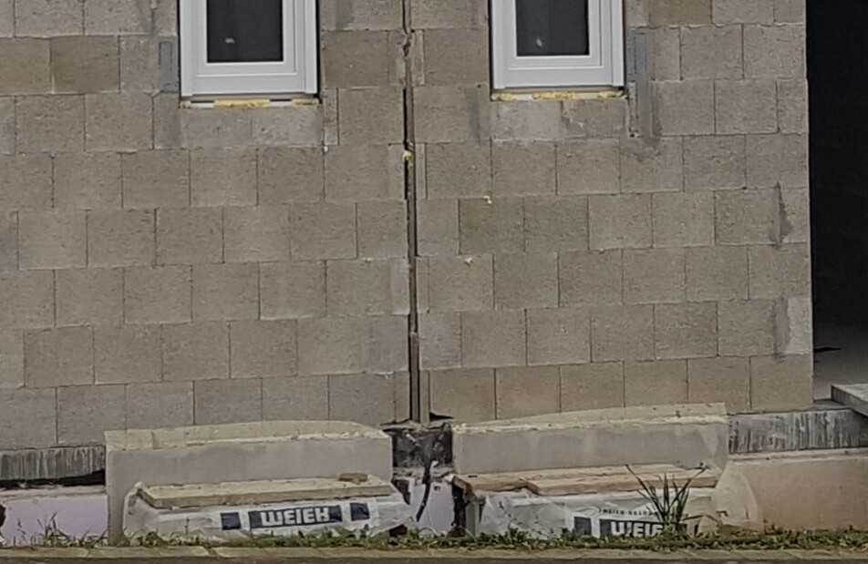 Außenwand aus Betonsteinen mit senkrechtem Riss zwischen zwei Fenstern; Baumaterialien am Boden.
