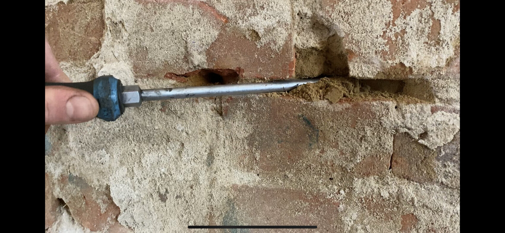 Person hält einen Metallstab durch ein Loch in einer bröckeligen Ziegelwand; Staub liegt am Mörtel.