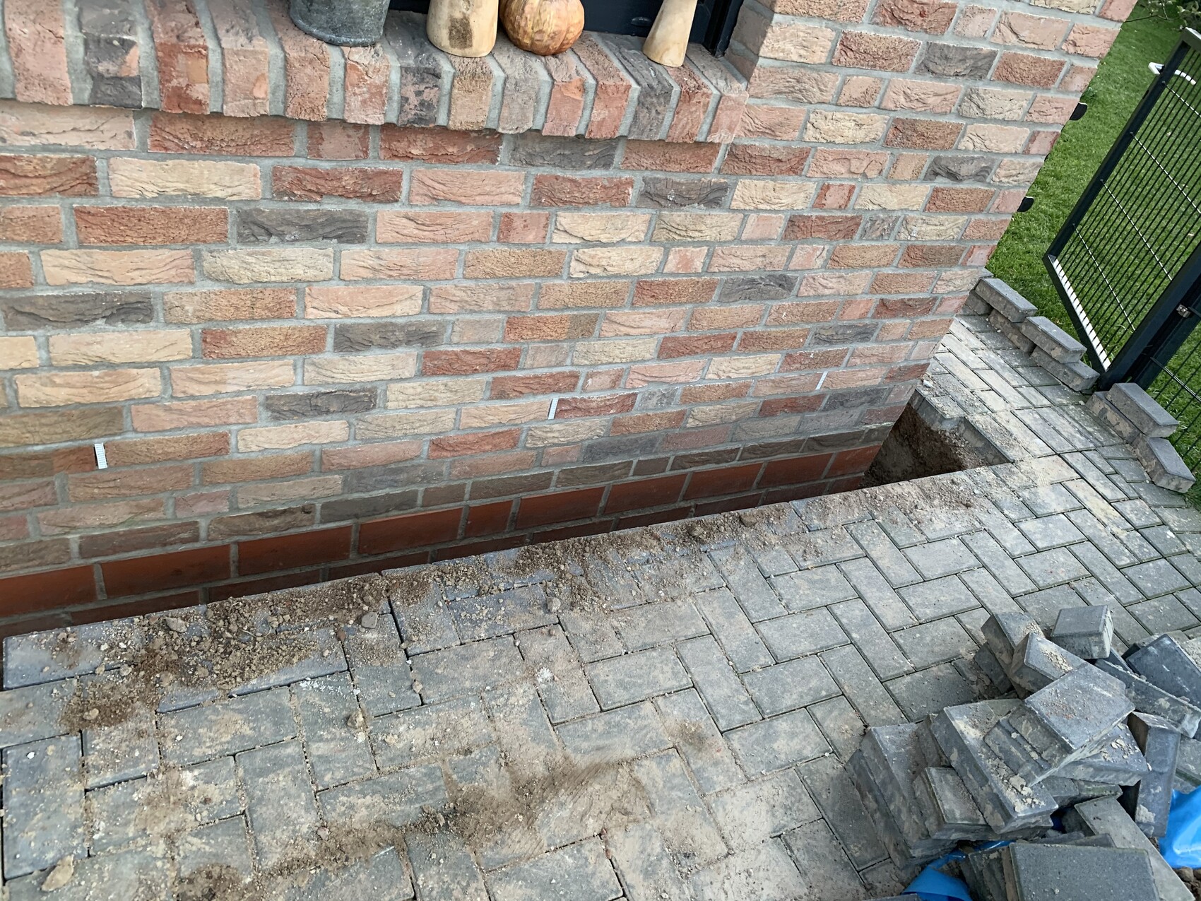 Backsteinwand mit offener Baugrube neben Terrasse, gepflasterter Boden, Baustellenschutt.