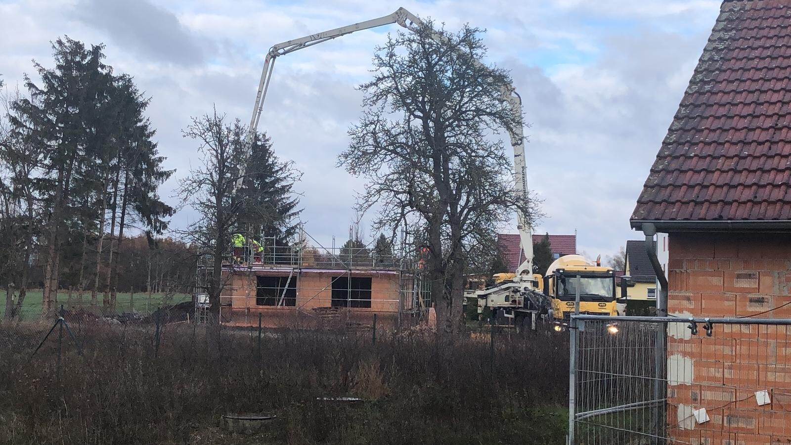 Baustelle: Betonpumpe mit langem Arm gießt Beton auf Rohbau; Arbeiter in Warnkleidung auf dem Gerüst.