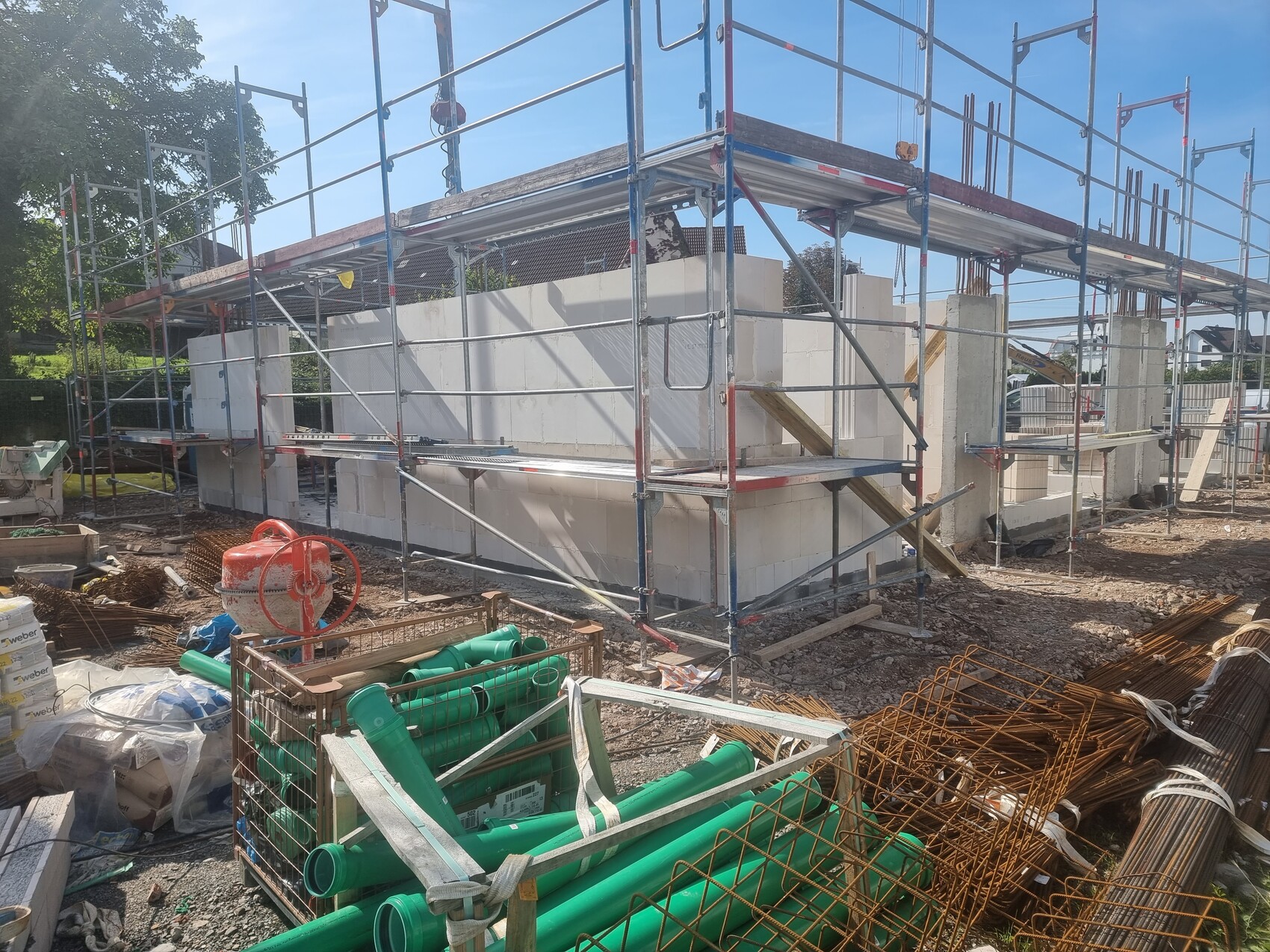 Baustellengerüst um Rohbau aus Betonblöcken; grüne Rohre und Baumaterialien im Vordergrund.