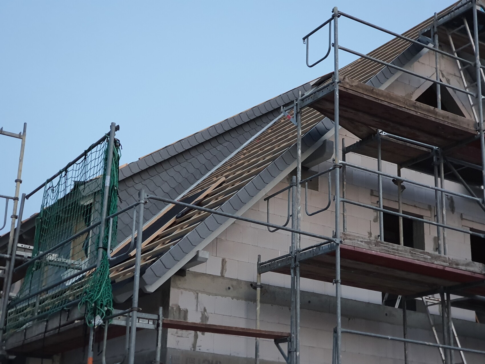 Baustelle: Gerüst um das Haus, Dach mit grauen Ziegeln, Holzlatten und grüne Netze.