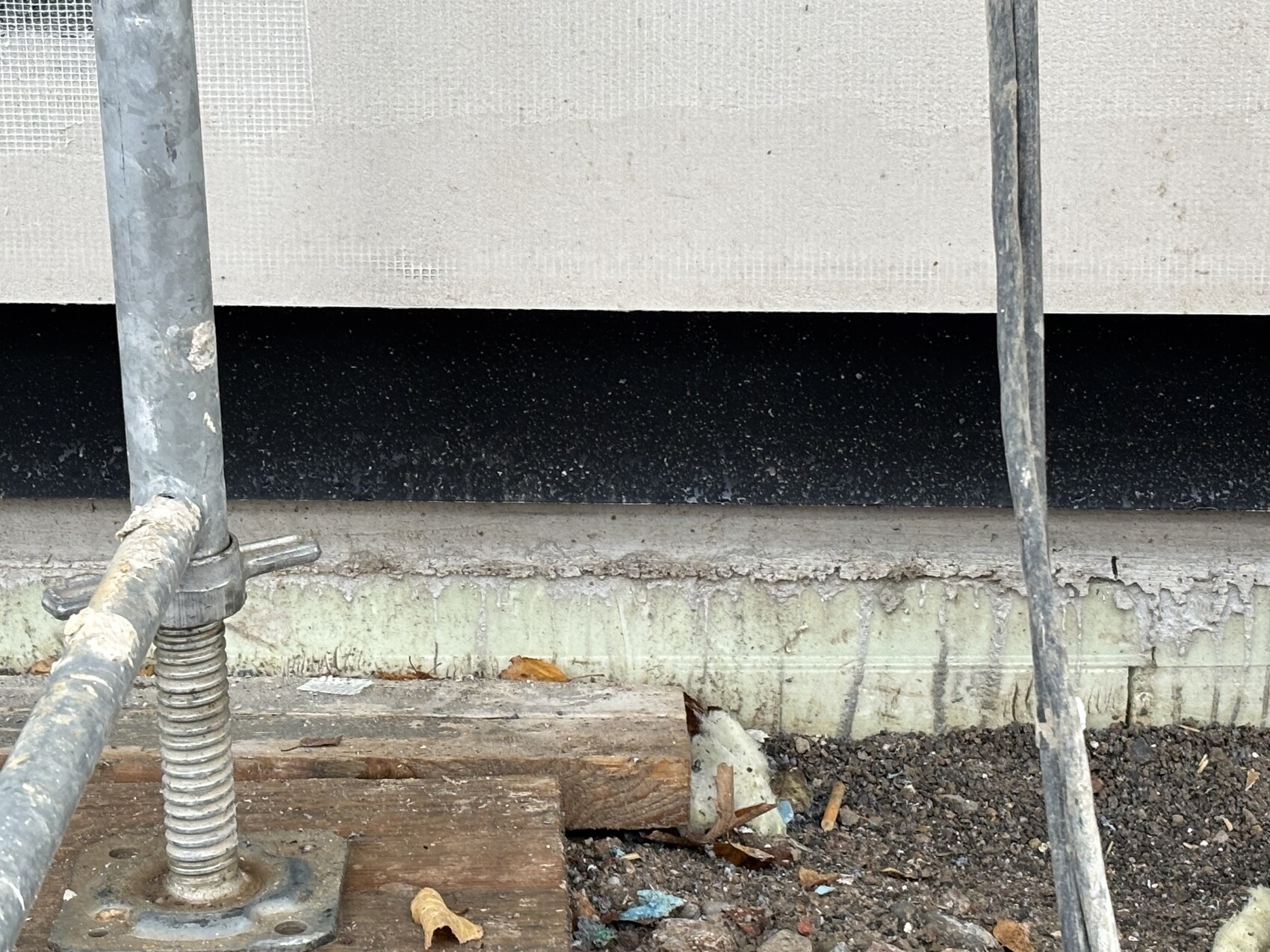 Metallgerüst-Stange mit Schraubfuß auf Baugrund, daneben Schutt und Holzreste vor Wand.