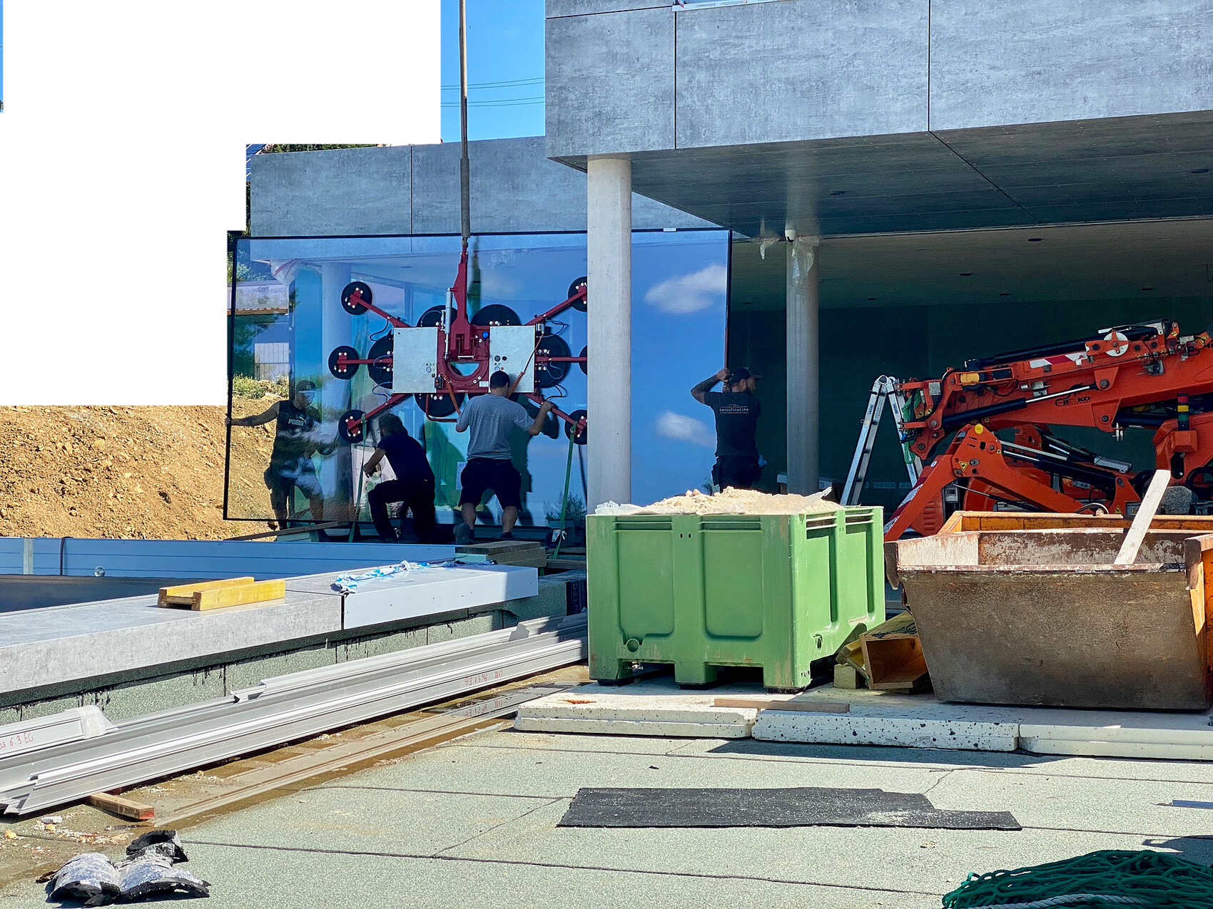 Bauarbeiter montieren eine Glasfassade; vorne stehen grüne Container und Bauabfälle auf der Baustelle