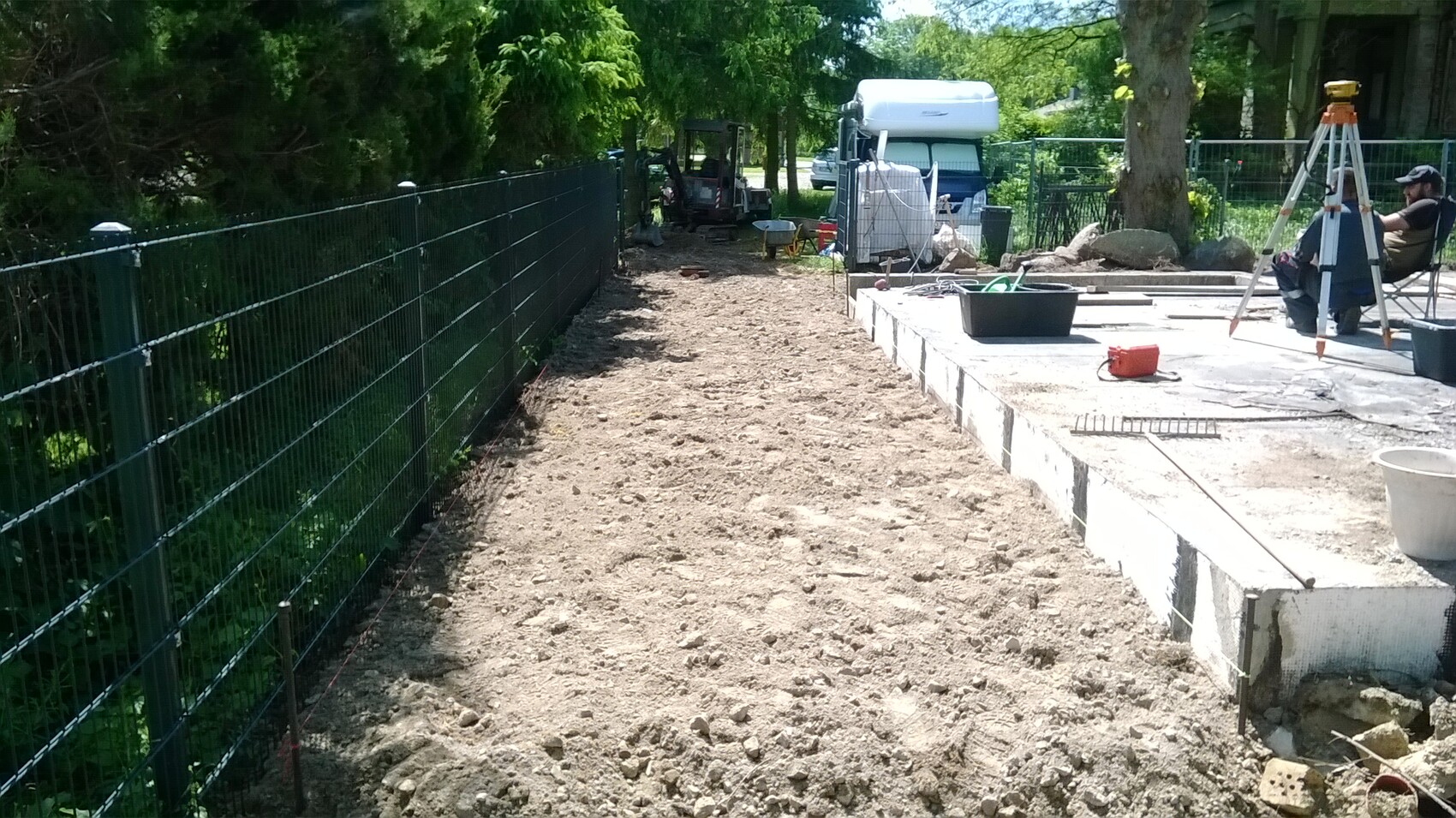 Garten-Baustelle: Kiesweg neben Metallzaun; Betonplatte rechts, Werkzeuge, Mann am Stativ.