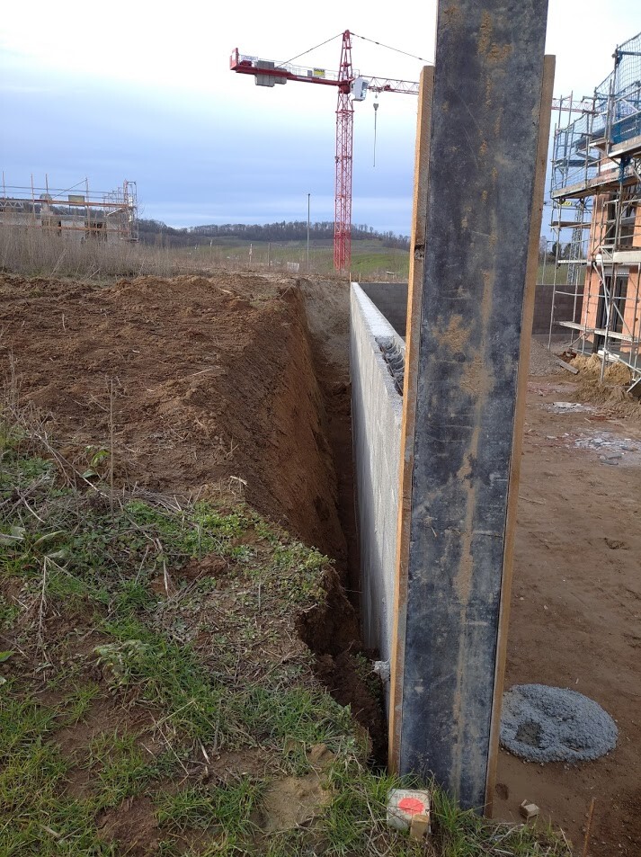 Tiefe Baugrube neben Betonwand, Stahlträger, Kran und Gerüst auf einer Baustelle.