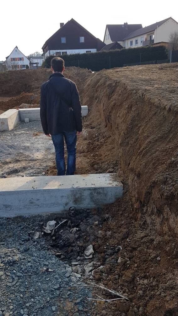 Mann von hinten in dunkler Jacke vor Baugrube mit Betonfundament und Erde.