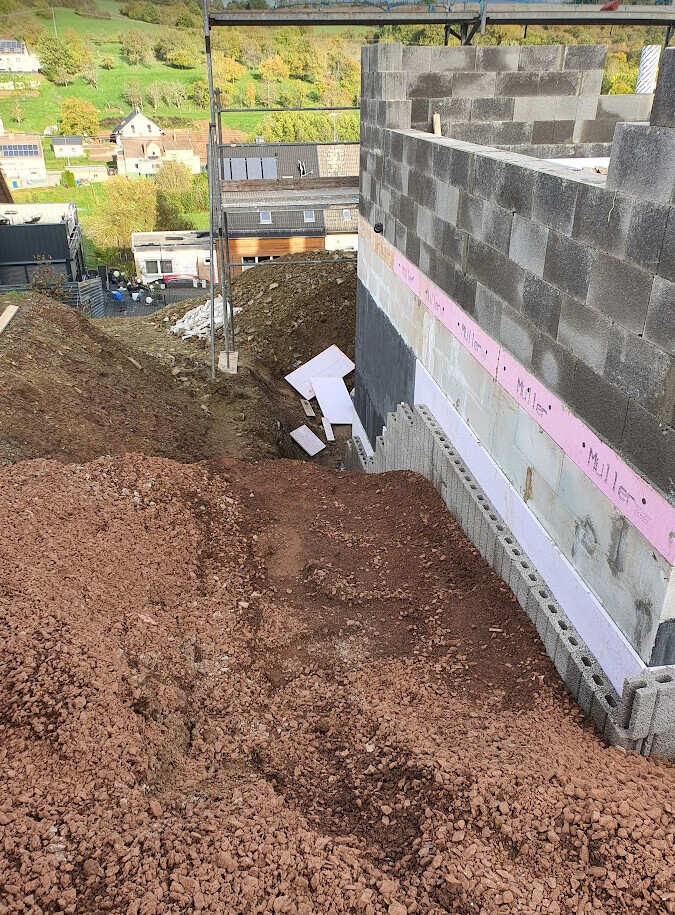 Baustelle mit unfertiger Betonblockmauer, Erdwall und Baugerüst; Hintergrund: Häuser am Hang.