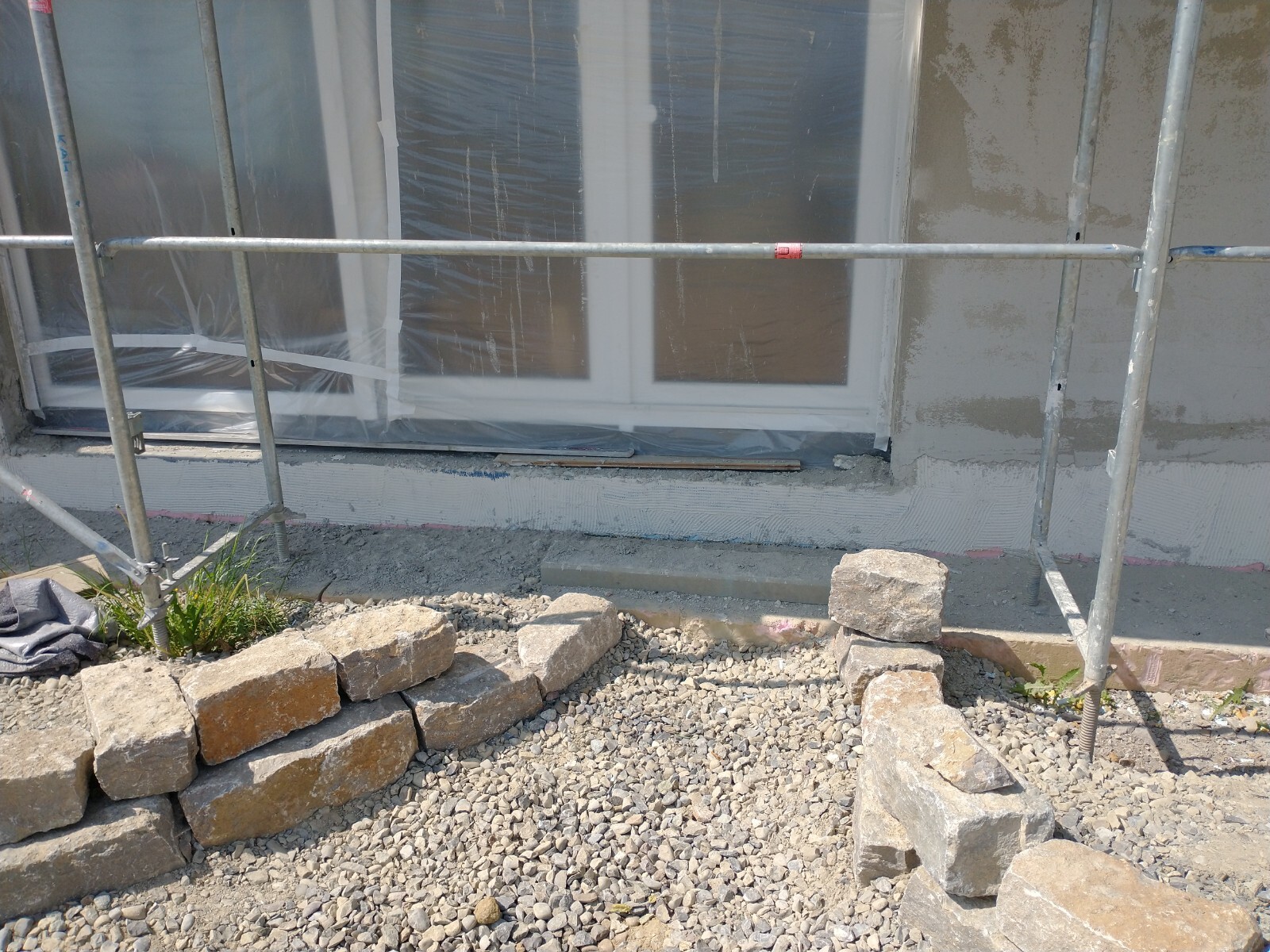 Baustelle mit Gerüst, Fensterabdeckung aus Plastik, Kiesboden und Steinmauer.