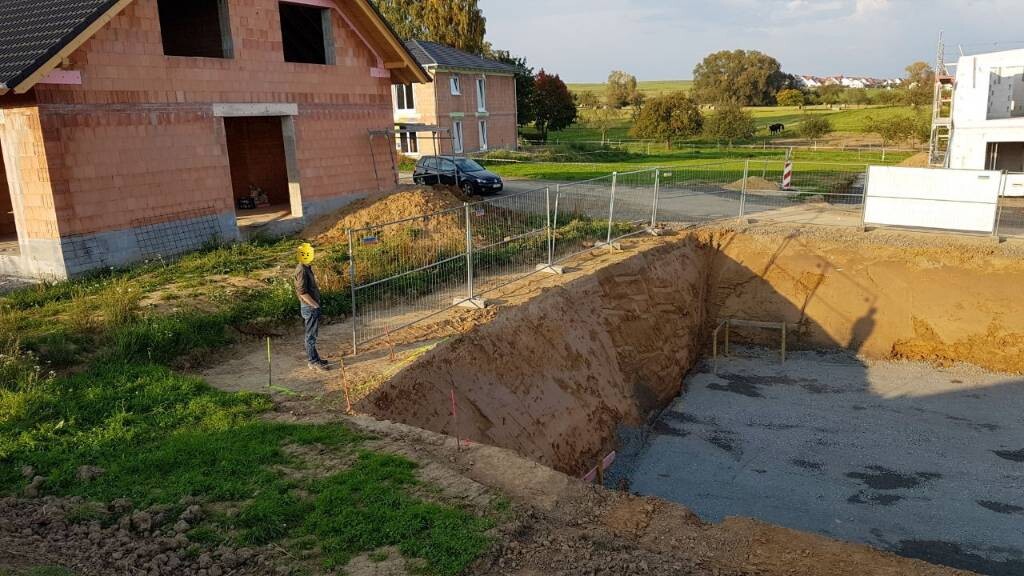 Baugrube mit Zaun, Arbeiter mit Helm an der Baustelle, Häuser im Hintergrund
