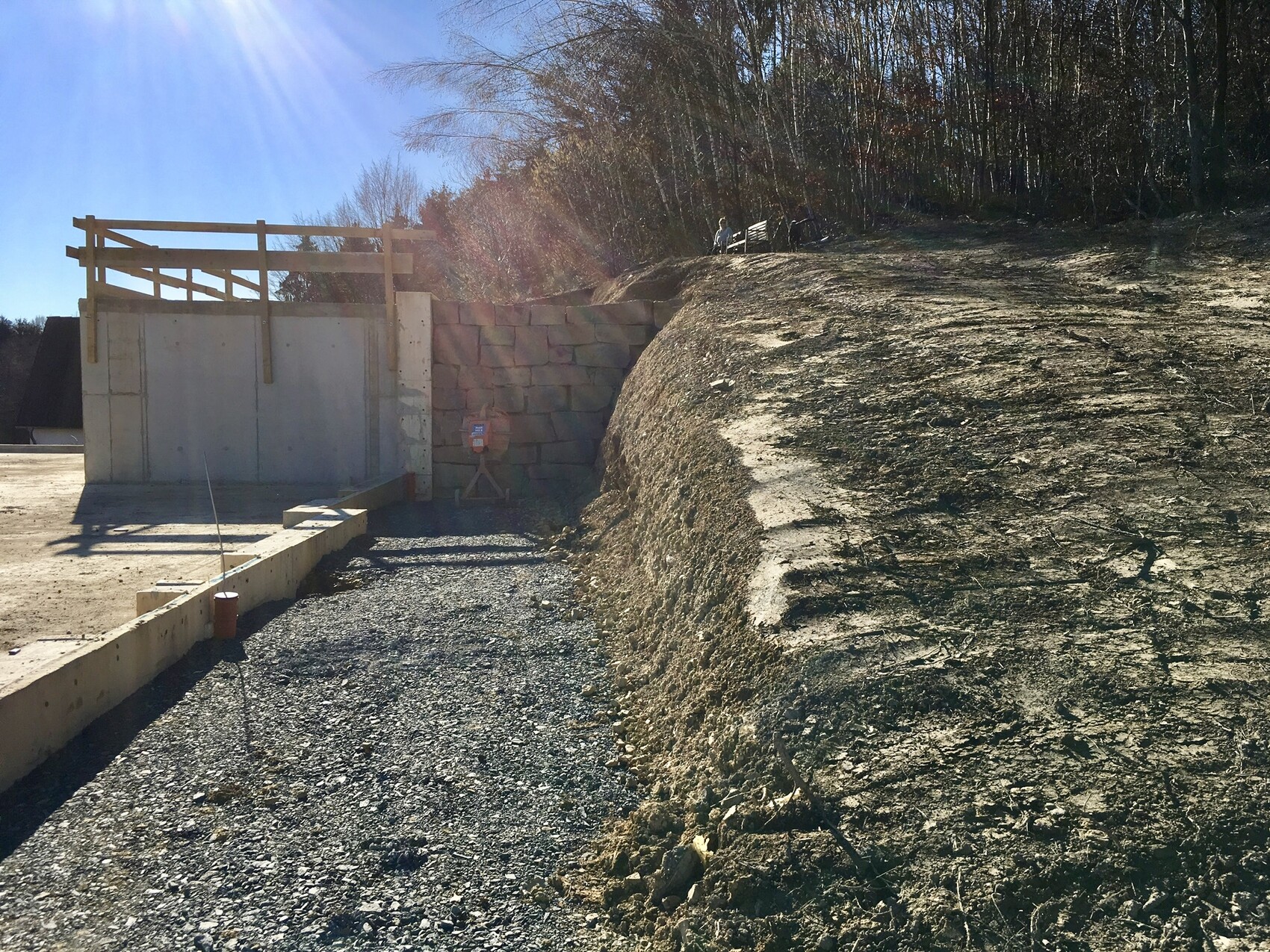 Baustelle mit Betonwand, Holzgeländer, Kiesweg und Erdwall, Bäume dahinter.