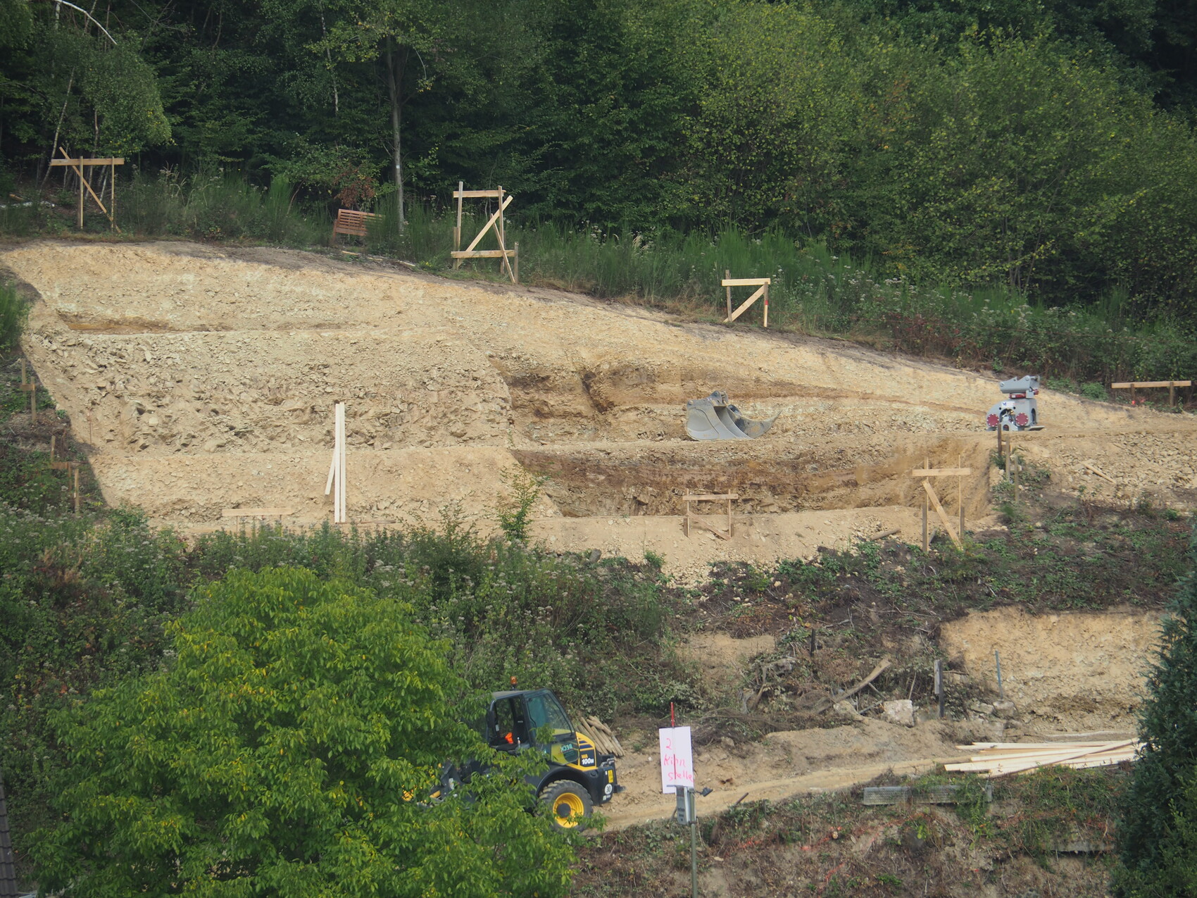 Hangkante mit abgetragenem Erdreich, Holzkonstruktionen, Bagger-Schaufel und Wald im Hintergrund.