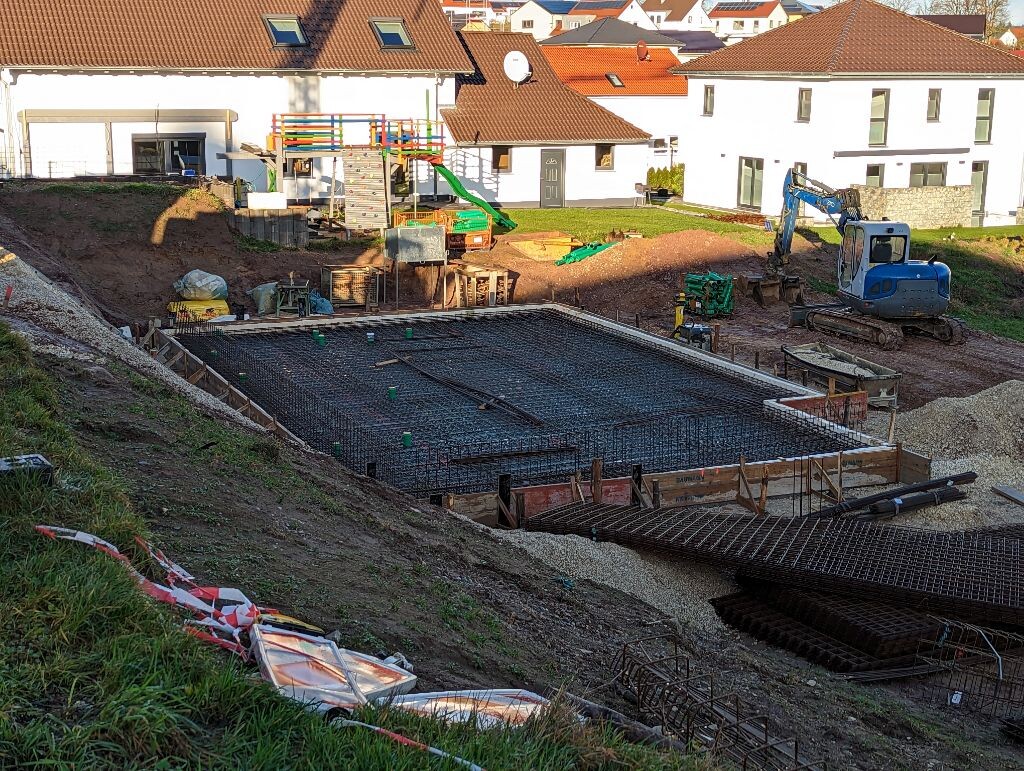 Baustellenfundament mit Stahlbewehrung, Holzformen, Bagger und Wohnhäuser dahinter.