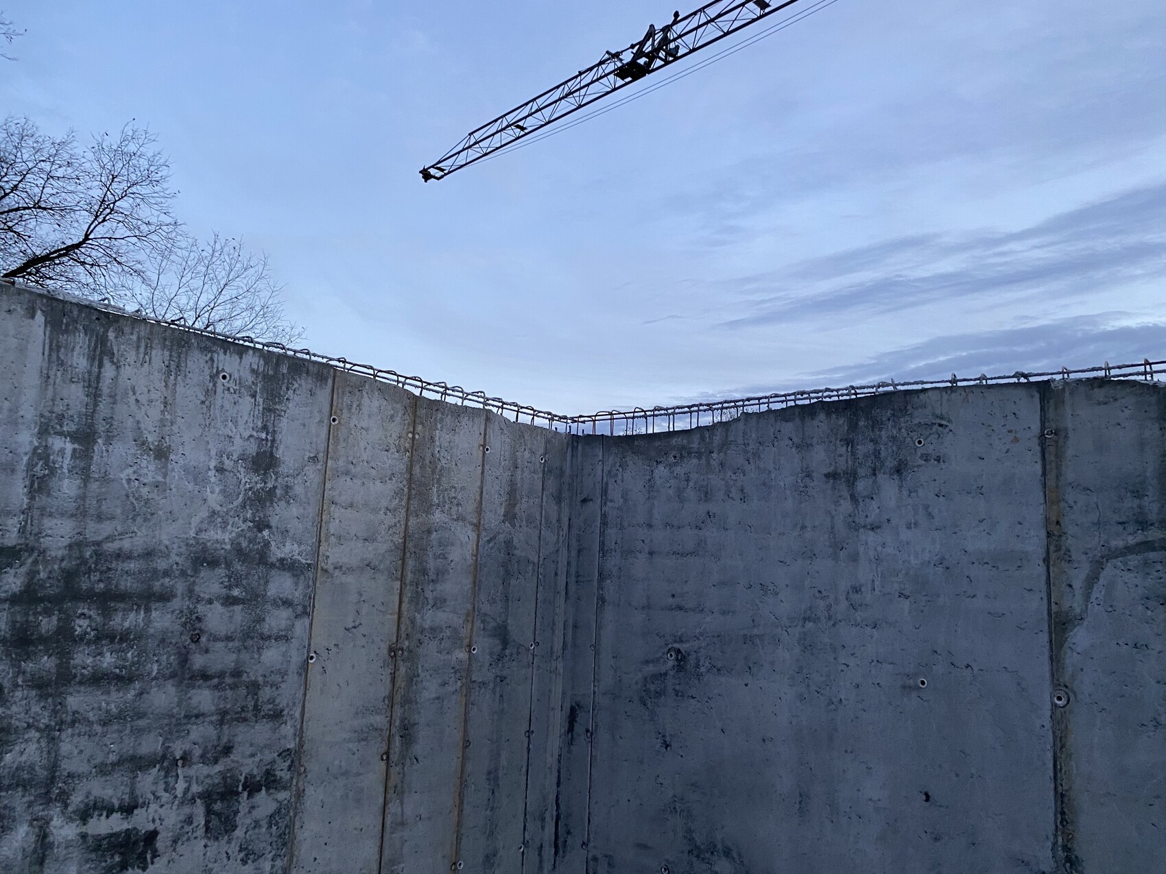 Baugrube mit hohen Betonwänden, Stacheldraht am Rand, Kran im Himmel über der Baustelle