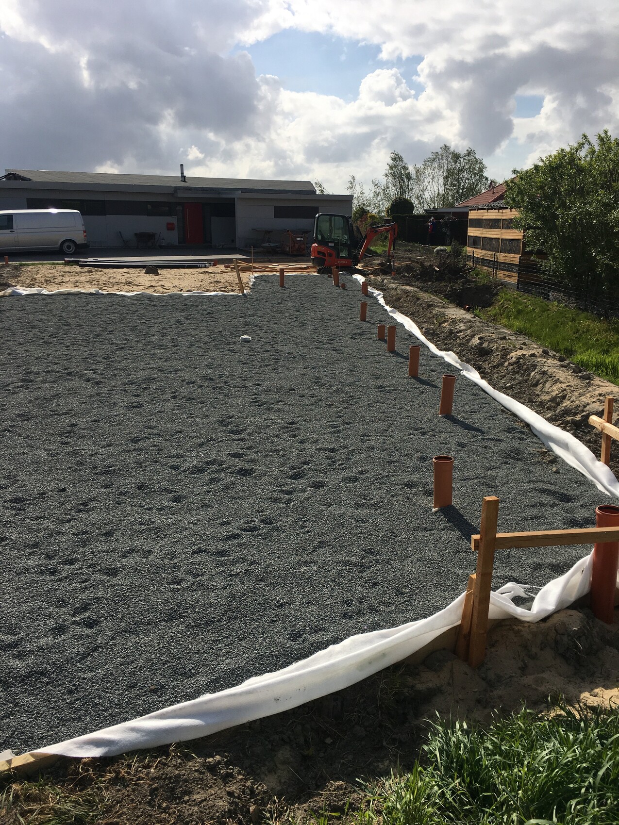 Baustelle mit Kiesunterlage, orange Rohre und Bagger, Geotextil, Haus im Hintergrund.