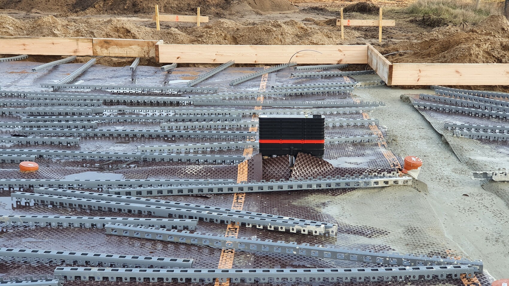 Baustelle: Fundamentboden mit Metallträgern, Holzformrahmen, orange Deckel, schwarzer Werkzeugkasten.