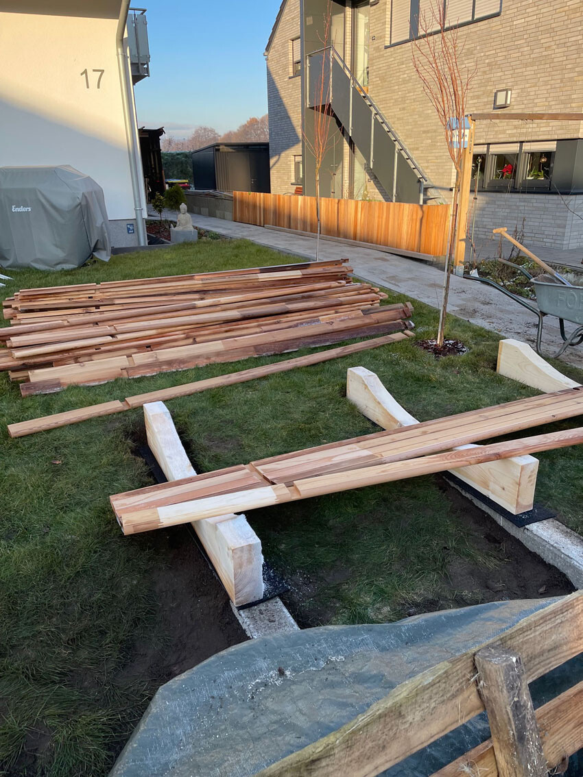 Baustelle im Innenhof: Holzplanken gestapelt, provisorische Brücke über Graben, Schubkarren rechts.