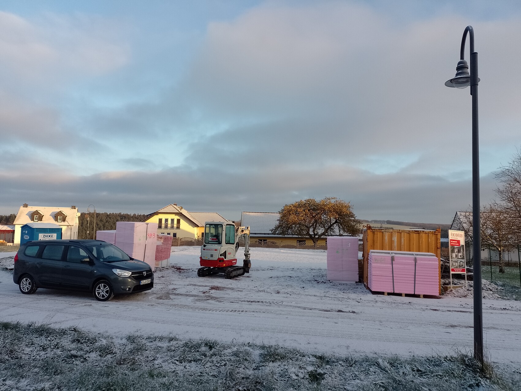 Baustelle mit Kleinstbagger, Dämmplatten-Paletten, Pkw und Laterne im Schnee