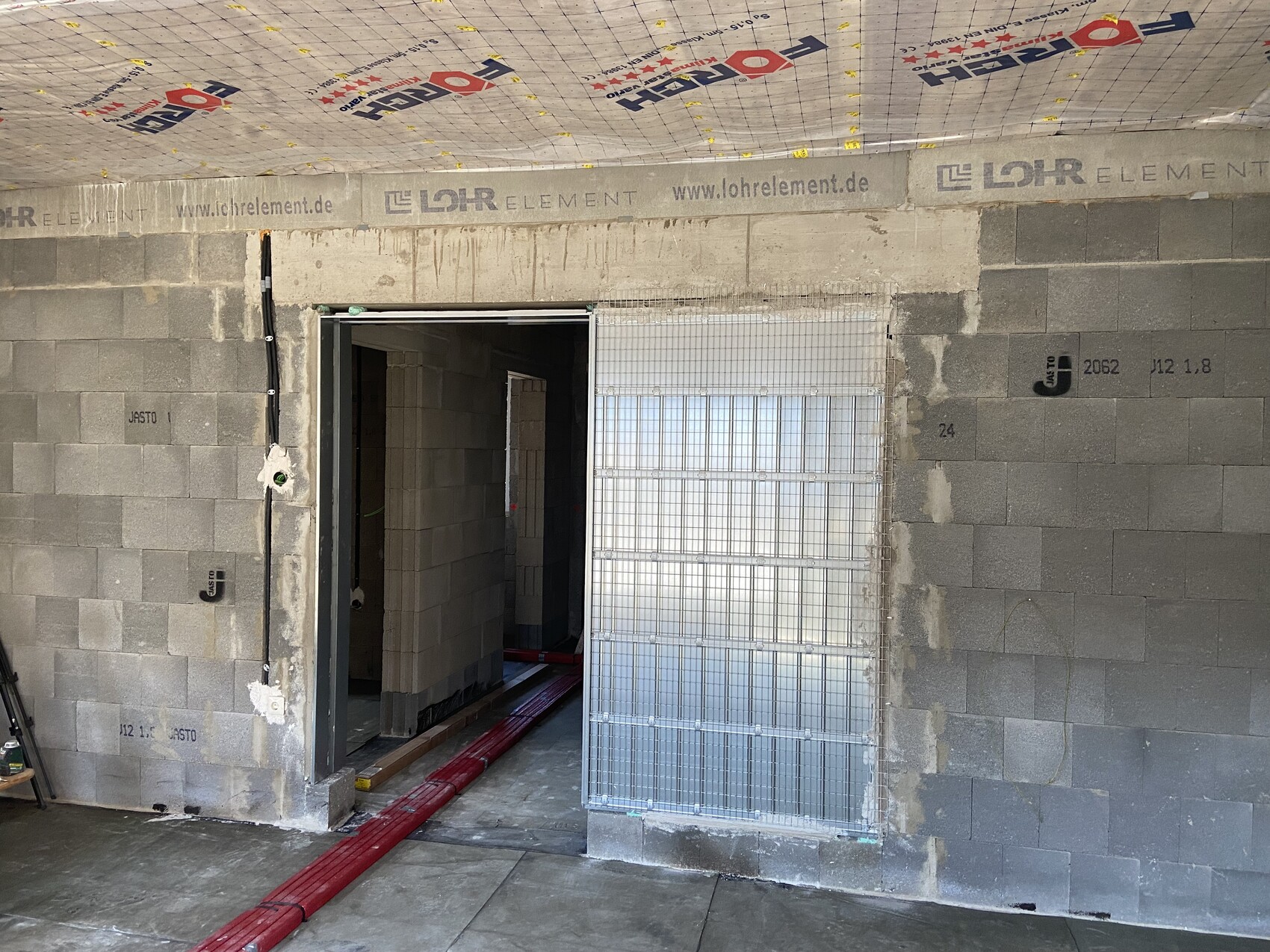 Baustelle: Innenraum mit Betonblöcken, Türdurchgang, rote Rohre am Boden, Metallgitterfenster.
