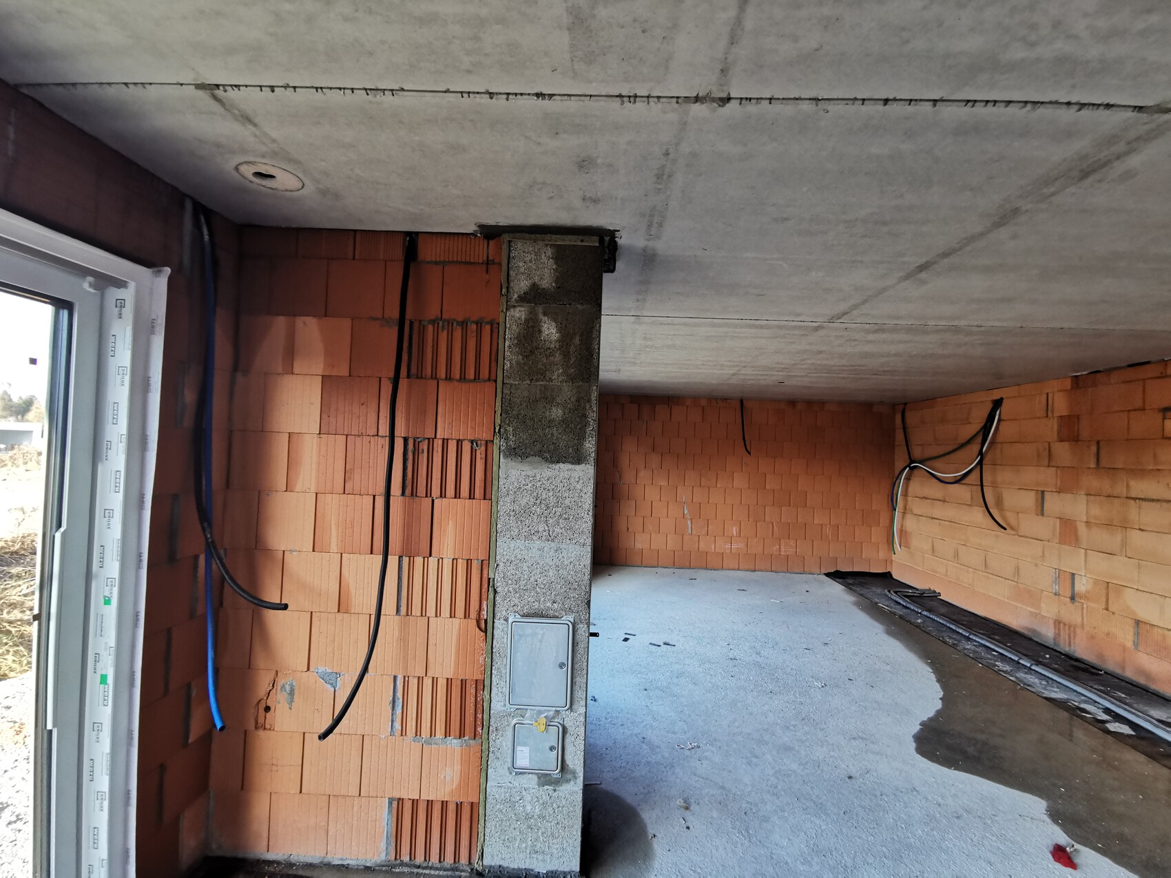 Unfertiger Innenraum mit Ziegelwänden, Beton-Decke, losen Kabeln und Wasserpfütze auf dem Boden.