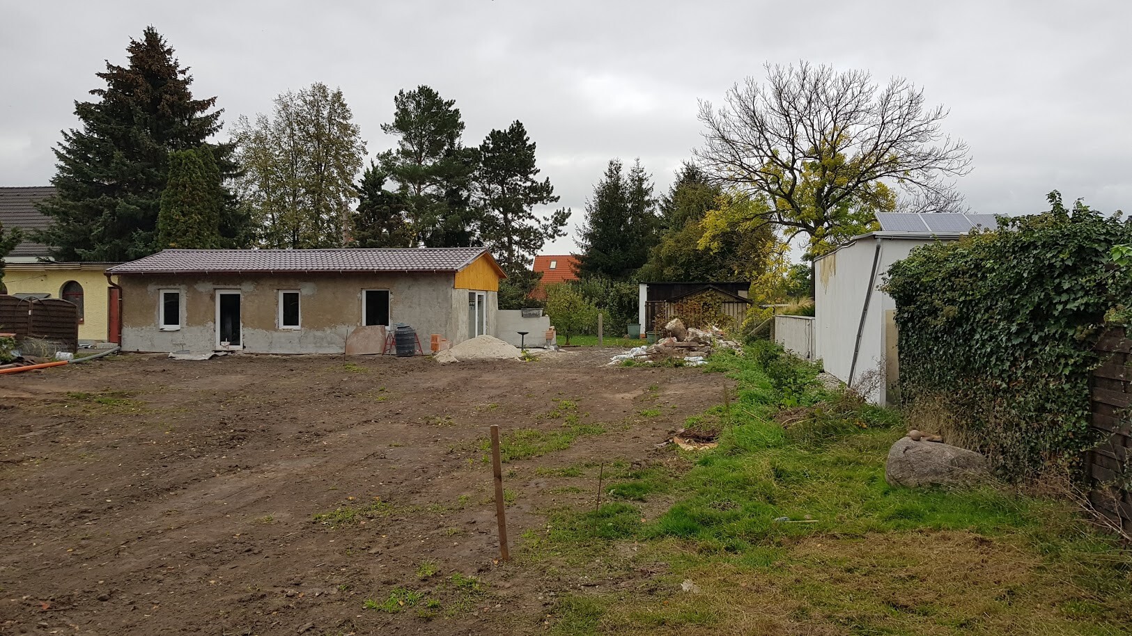 Unfertiges Baugebäude auf einer Baustelle mit Erde, Sandhaufen und Bäumen dahinter.