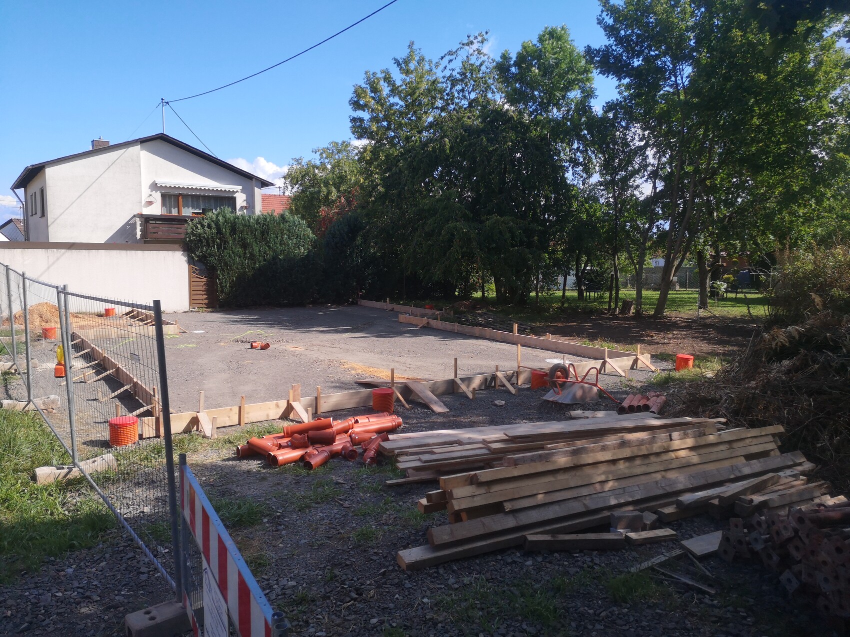 Baustelle im Garten: Holzformen, rotes Rohr, Holzstapel, Kiesboden, Haus links, Bäume rechts.