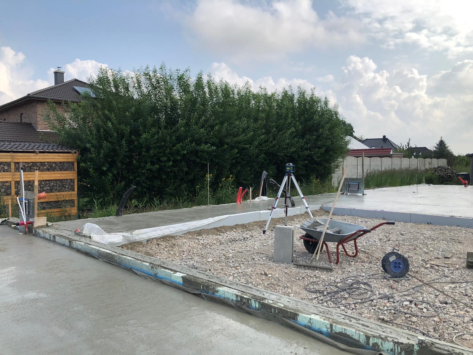 Baustelle mit Fundamentplatten, Lasermessgerät auf Stativ, Schubkarren, Werkzeuge, Hecke dahinter.