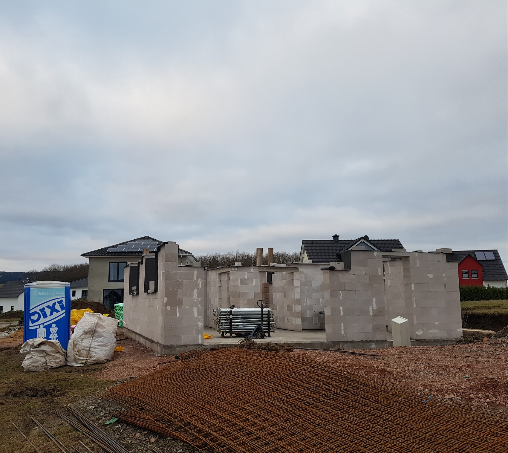 Baustelle mit Rohbau aus Betonblöcken, Bewehrung, Baumaterialien und Wohnhäusern im Hintergrund