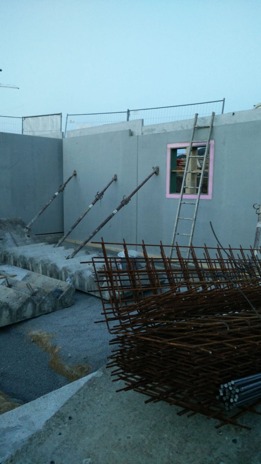 Baustelle: graue Betonwand, pinker Fensterrahmen, Leiter, Bewehrung und Staub im Vordergrund.