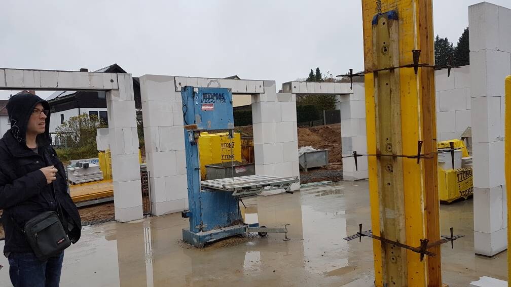 Person im Hoodie vor einer Baustelle mit weißem Mauerwerk, blauer Baumaschine und nasser Fläche.