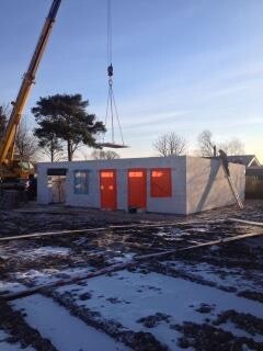 Rohbau-Baustelle mit Kran hebt Wandbauteile, kleinstes Gebäude mit orange Türen im Schnee