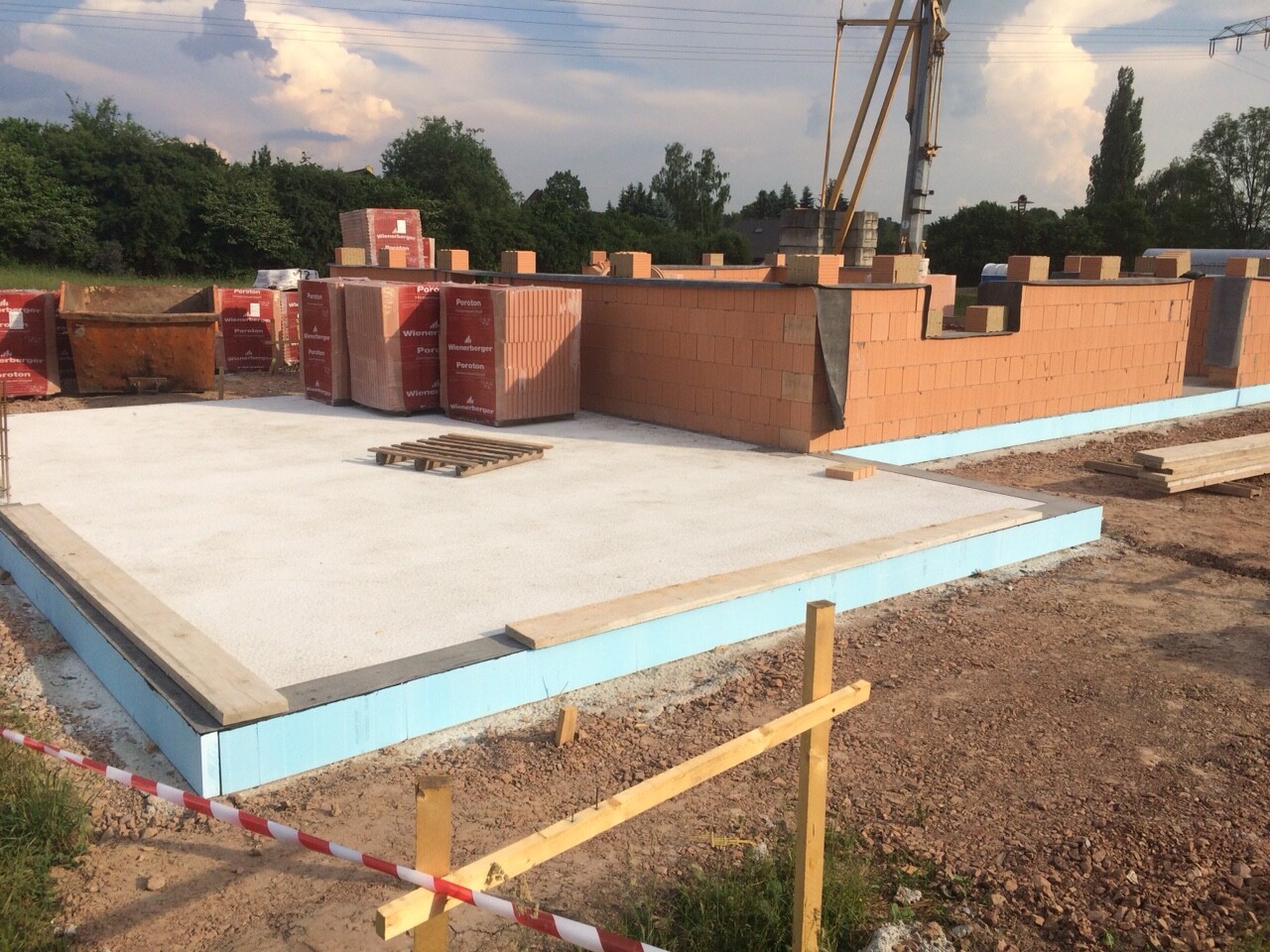 Baustelle: Fundamentplatte, blaue Dämmung, rote Ziegelwände, gestapelte Baumaterialien.