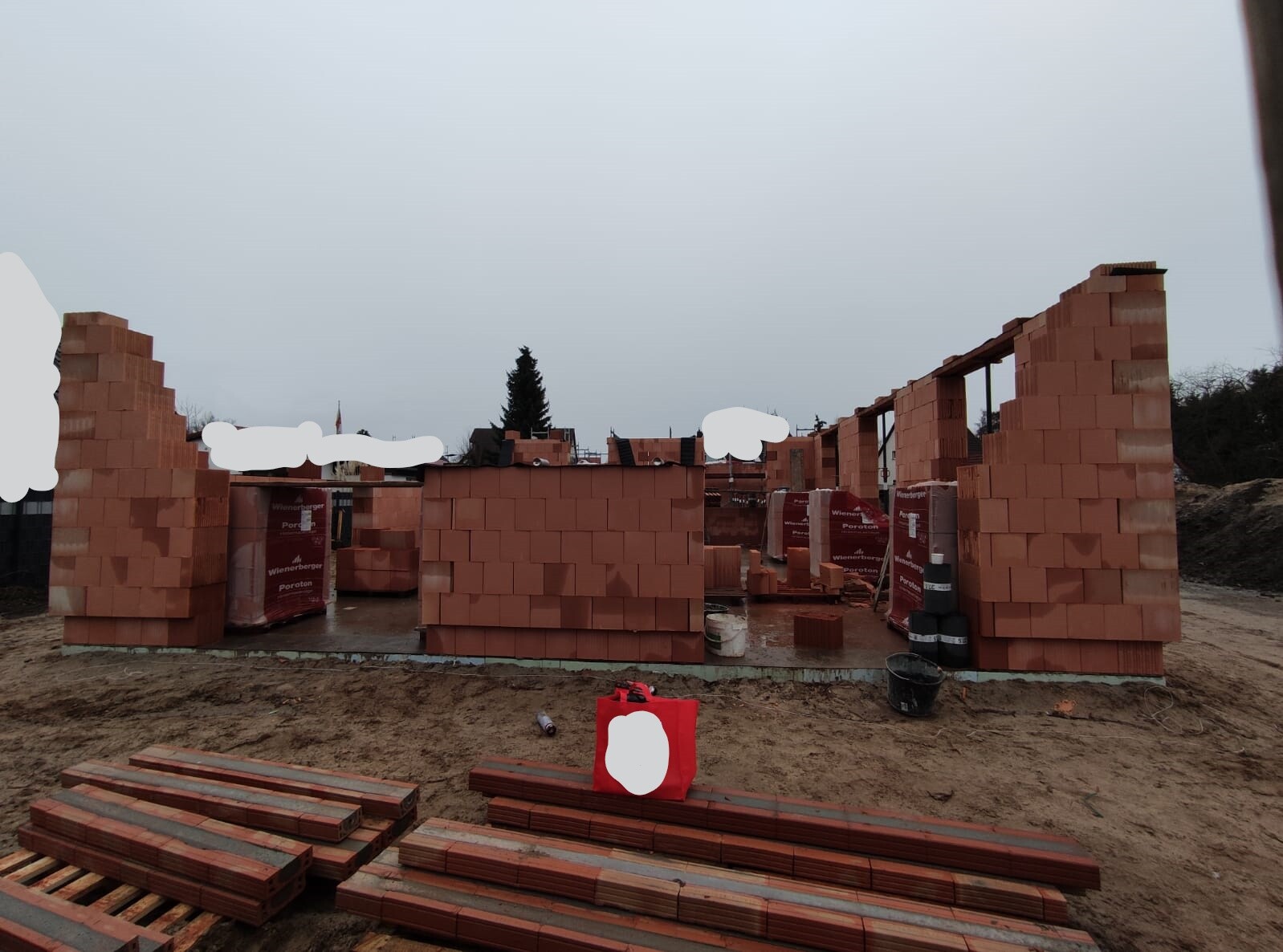 Unfertige Ziegelwände auf einer Baustelle, Stapel roter Ziegelsteine und Bauwerkzeuge sichtbar.