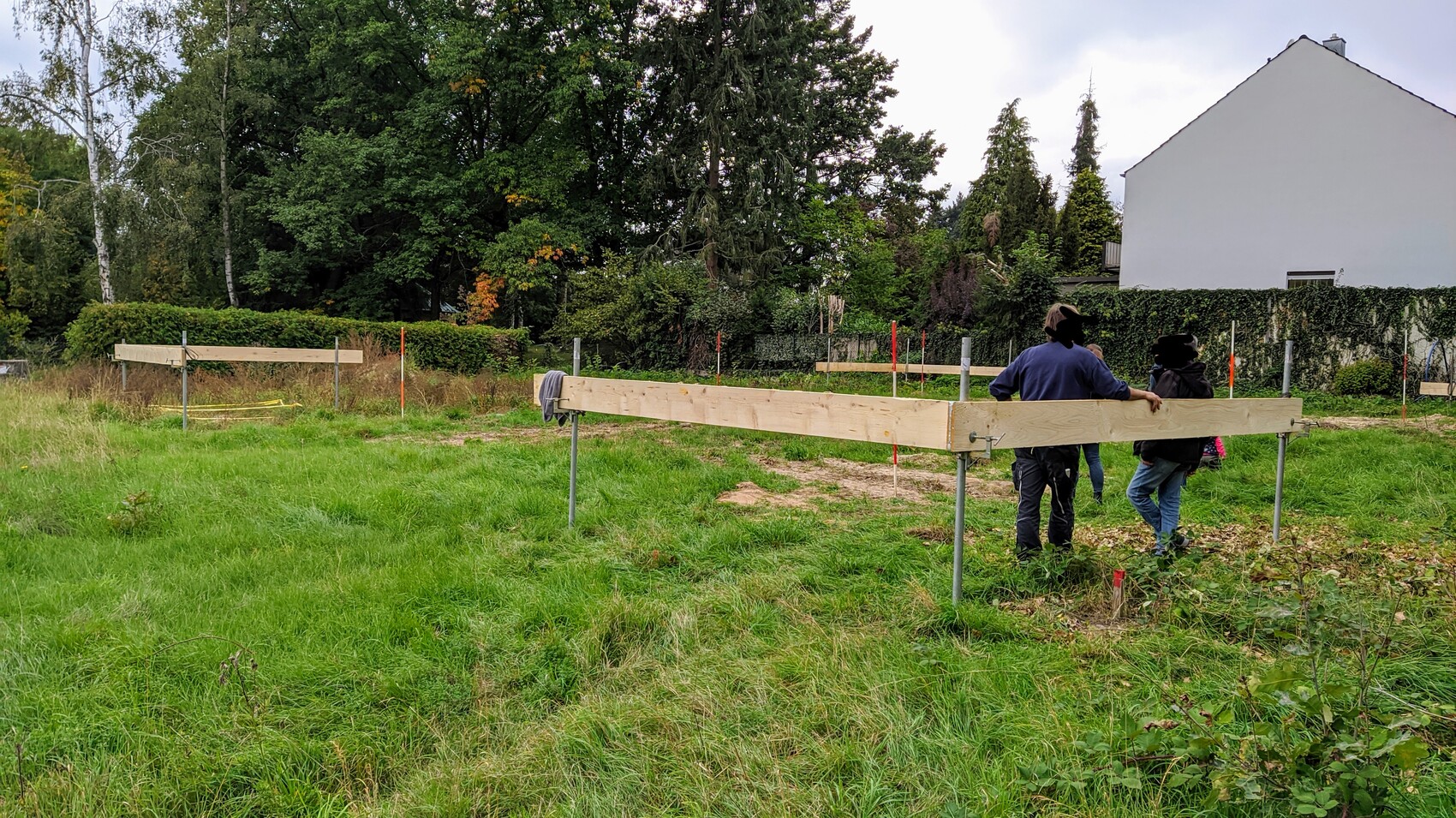 Grünes Feld mit Holzbalken auf Metallstützen; mehrere Personen stehen daneben, Bäume dahinter.