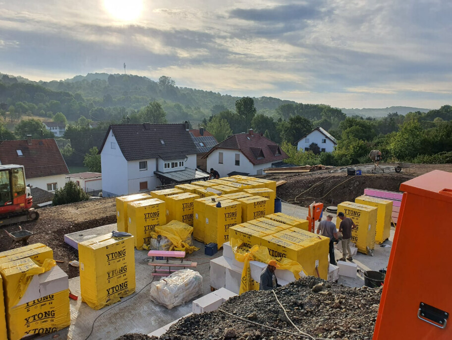 Baugelände mit gelben YTONG-Steinen, Bauarbeiter, Häuser am Hang, orange Baumaschine.