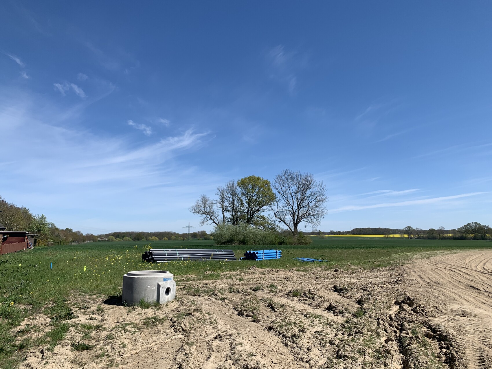 Offenes Feld mit gestapelten Rohren, runder Betonbehälter, blauer Himmel.