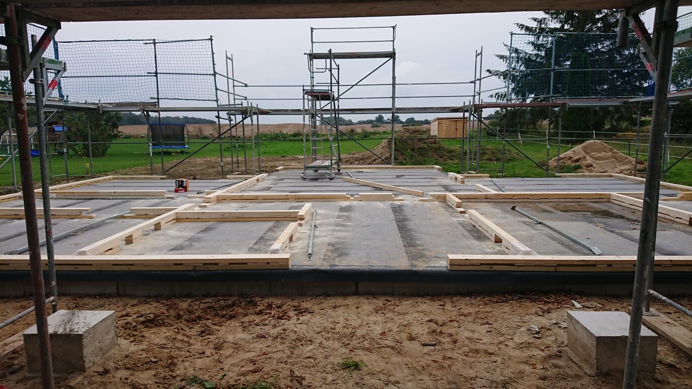 Baustelle: Holzrahmen auf Betonboden, umlaufendes Gerüst, offene Feldlandschaft im Hintergrund.