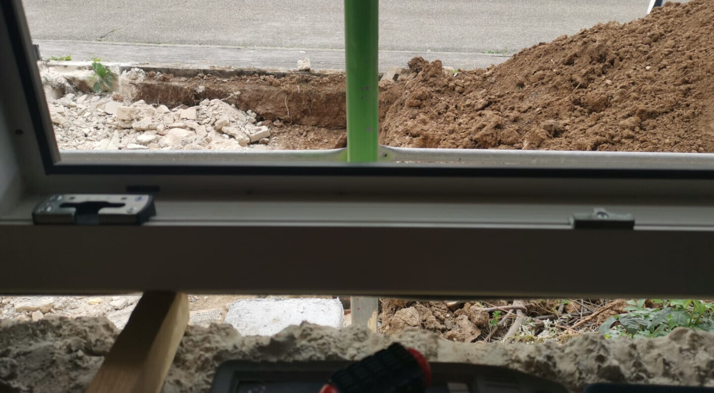 Blick durch Fenster auf Baustelle: Erde, Schutt, grüne Stange, Werkzeug im Vordergrund.