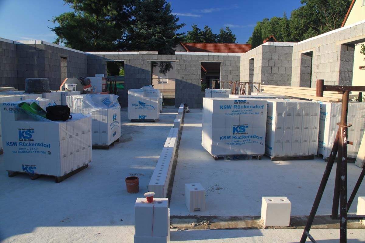 Baustelle mit gestapelten Betonblöcken auf Paletten und unfertigen Wänden.