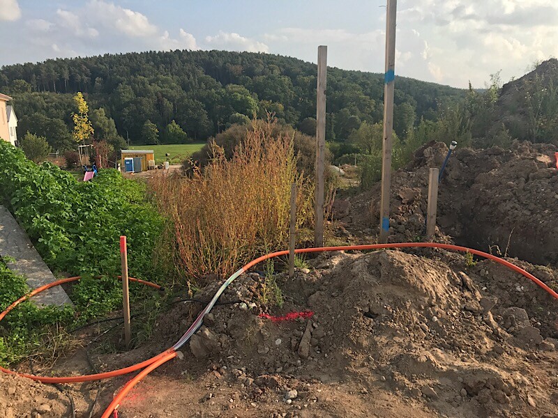Baustelle mit Erdarbeiten, orange Rohre, Holzpfosten und umliegende Wiesenlandschaft