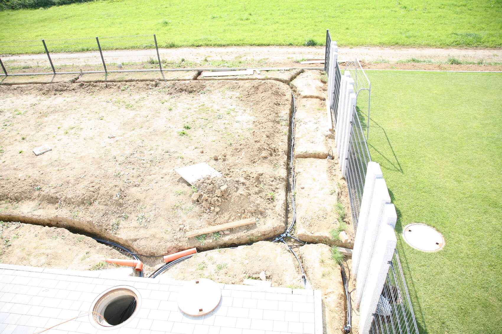 Ausgehobene Baustelle mit Gräben, Rohren, Fundamentarbeiten neben Gras und Zaun