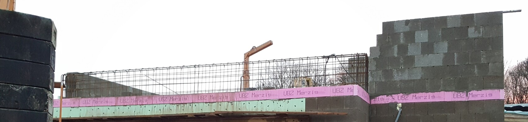 Rohbau: graue Betonblockwand, sichtbare Stahlbewehrung, pinke Dämmstreifen, linke Reifenstapel.
