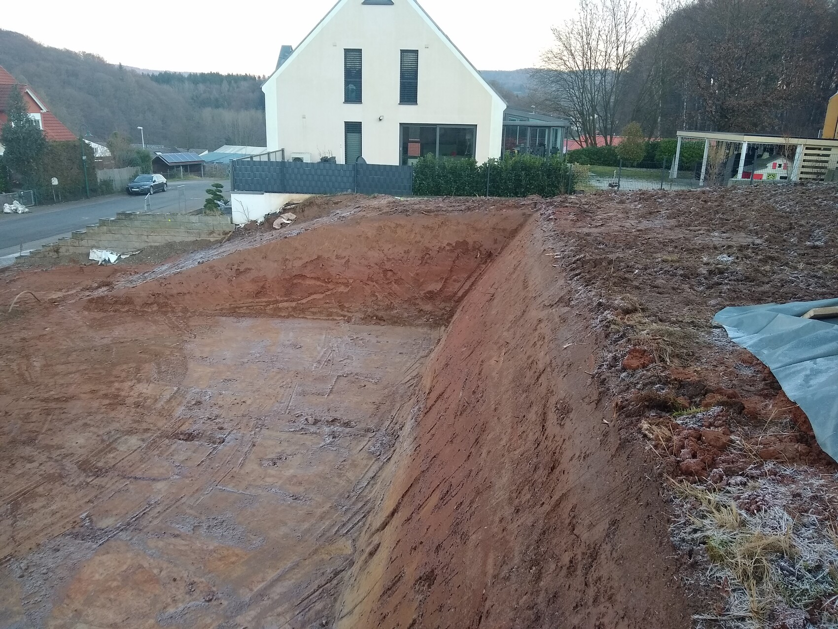Große Baugrube neben Wohnhaus; steile rote Erdwand, Gartenzäune, Straße und Auto im Hintergrund.