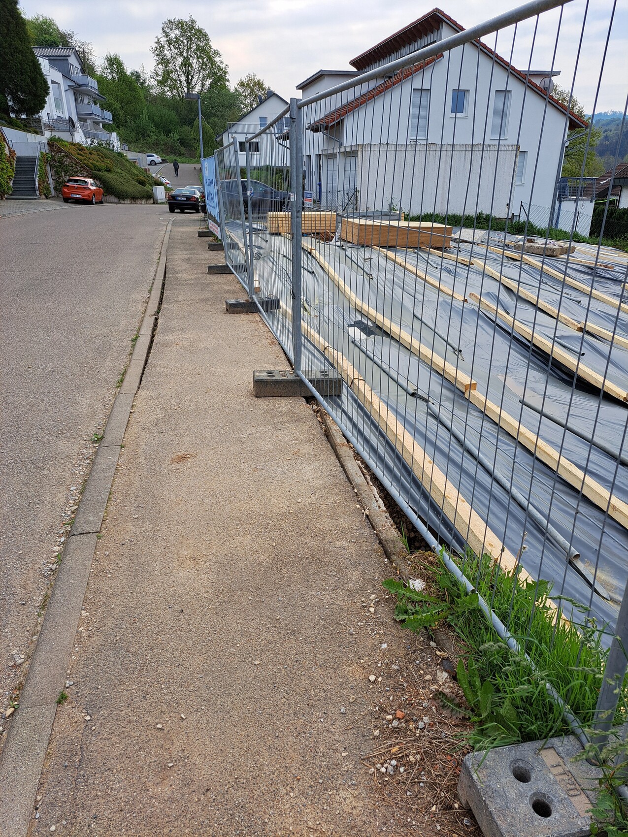 Baustelle am Straßenrand: Metallzaun, Bauholz und Folie, Häuser im Hintergrund.