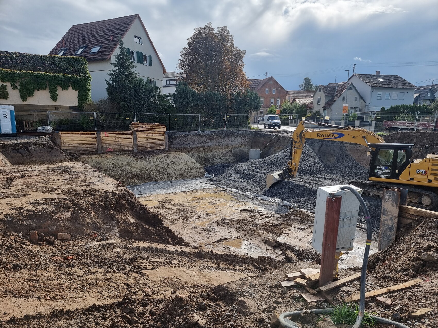 Gelber Bagger gräbt eine Baugrube in einer Wohnsiedlung; Kieshaufen und Holzbarrieren.