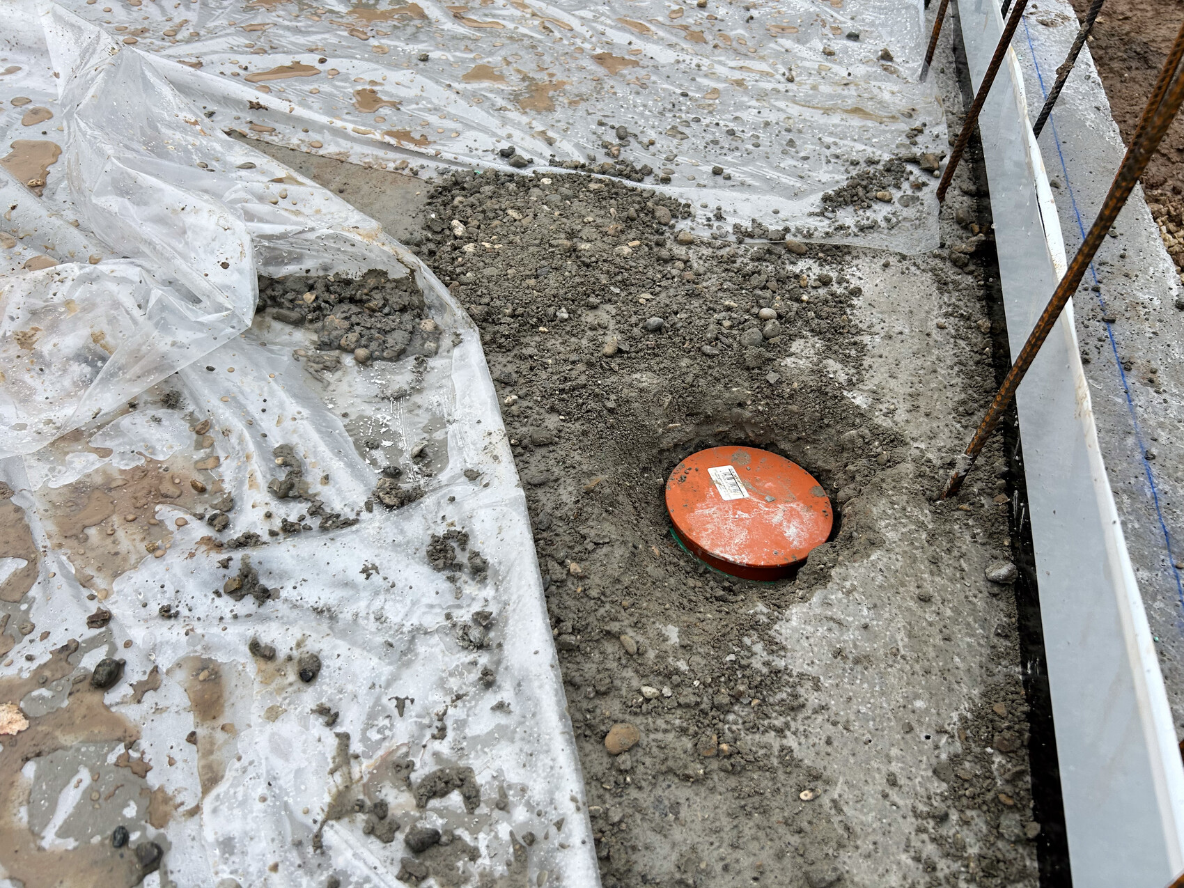 Baustelle: Erdarbeiten, Plastikfolie, Kies und roter runder Zugangdeckel im Fundamentbereich.