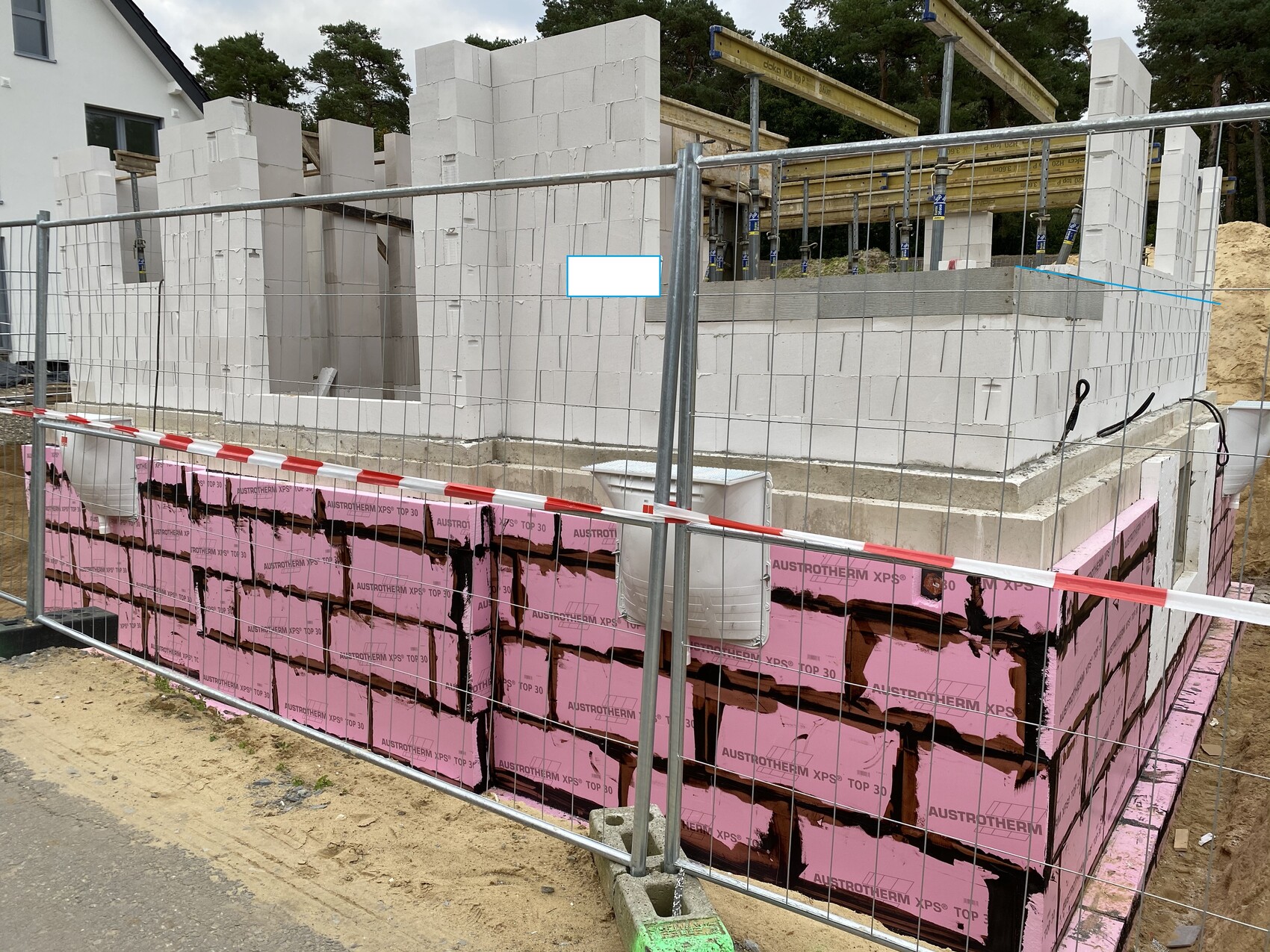 Baustelle mit weißen Betonblöcken, pinken Dämmplatten am Fundament, Metallzaun um das Gelände.