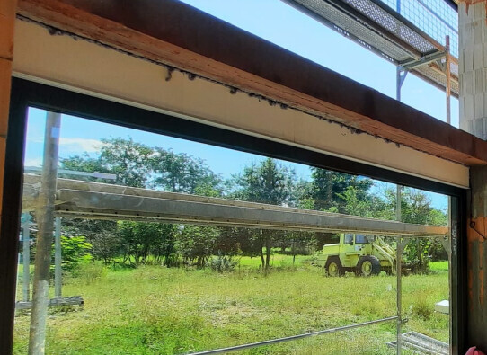 Durch ein offenes Fenster sichtbar: Gerüst, grüne Wiese und ein gelbes Baufahrzeug im Hintergrund.