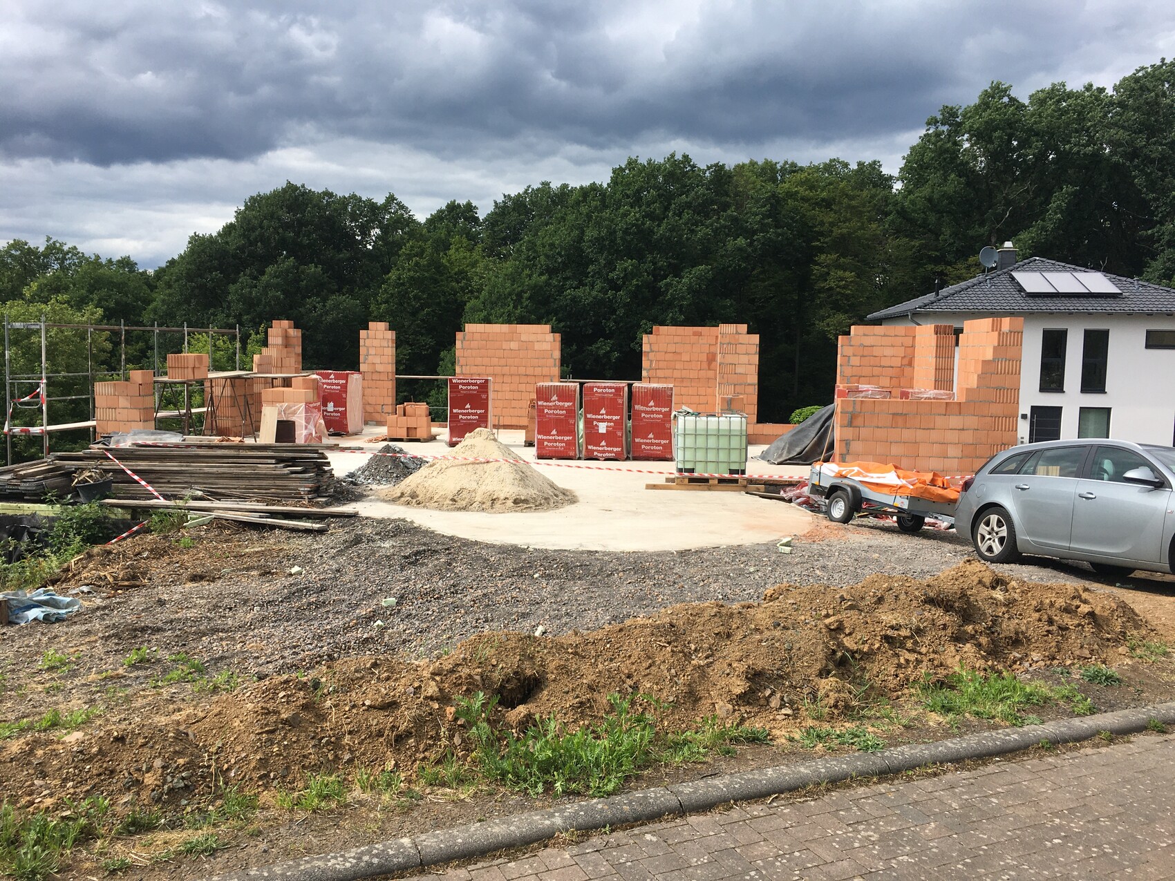 Baustelle mit unfertigen Ziegelwänden, Sandhaufen, Baustoffstapeln und PKW.